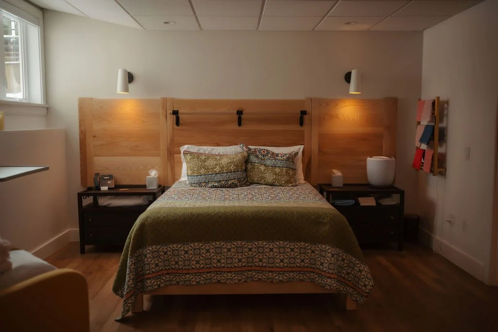 A cozy bedroom with a wooden headboard, two black nightstands with lamps, a patterned bedspread, and decorative pillows. There's a window on the left and a wall-mounted magazine rack on the right.