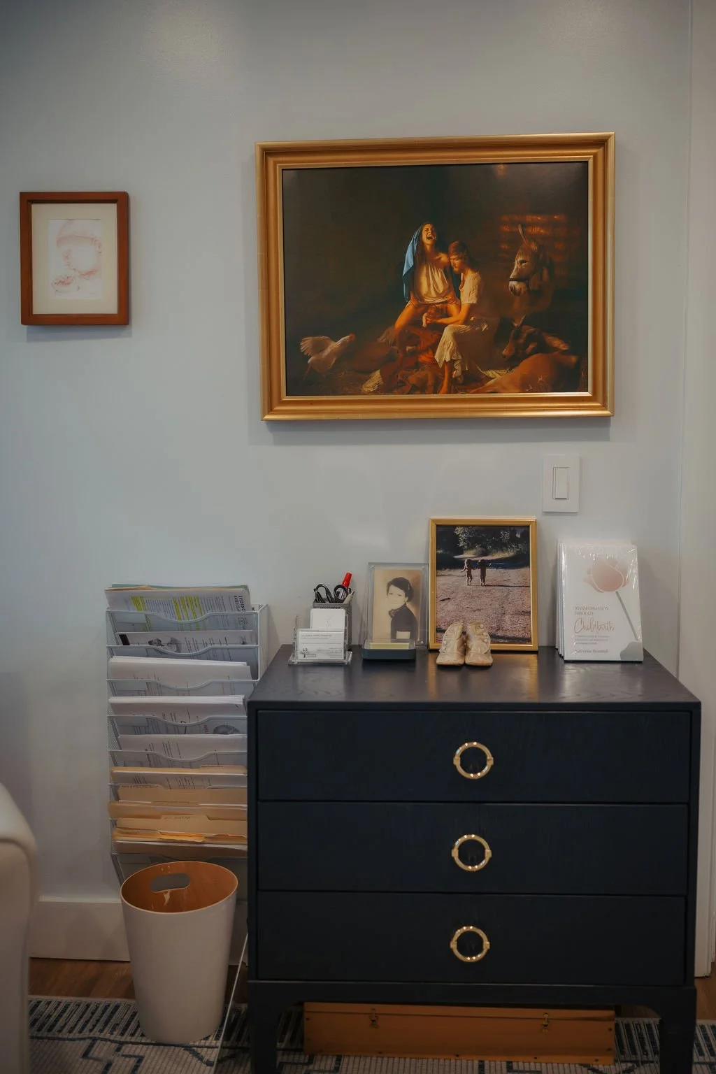 A black dresser with three drawers and gold ring handles, next to a white wire file organizer with papers, office supplies, framed photographs, a pair of shoes, and a tissue box on top. On the wall above, they are framed paintings and photos. The lar