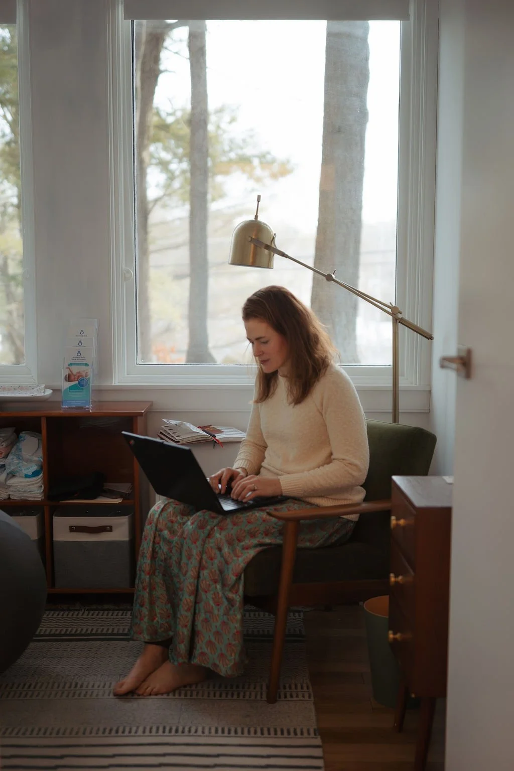 Woman sitting in a chair using a laptop in a cozy room, with a window behind her showing trees outside. There is a wooden shelf with items and a gold desk lamp over her head.
