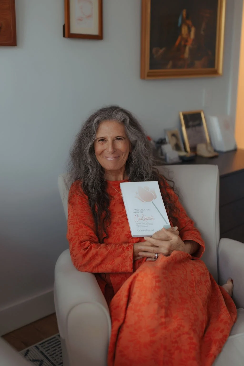 A woman with long gray hair smiling and sitting in a white armchair, holding a book with a floral cover in an indoor setting with framed pictures on the wall behind her.