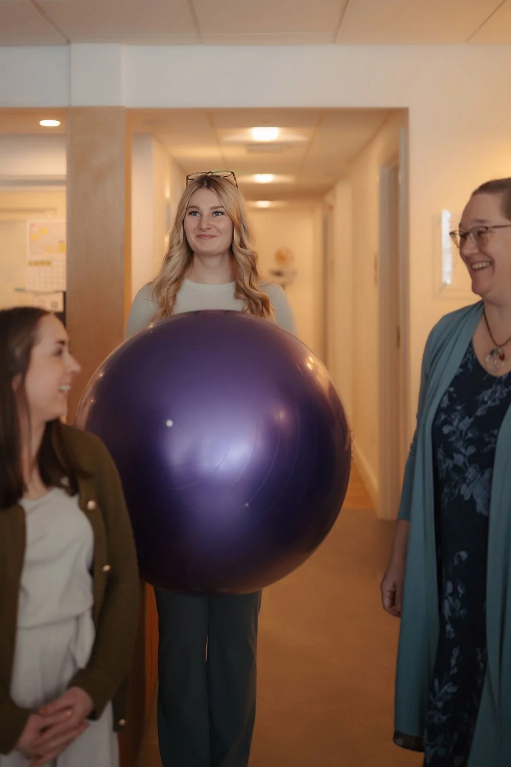 Four women standing in a hallway, with one holding a large purple exercise ball, smiling and engaging in conversation.
