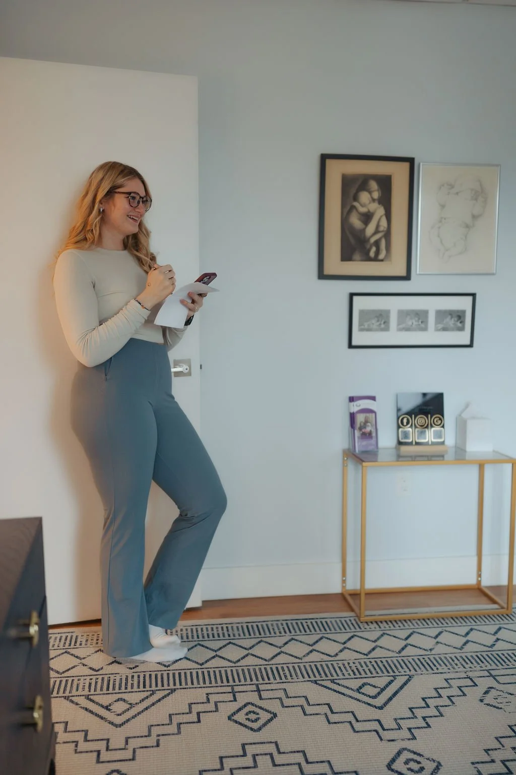 A woman with glasses and long blonde hair, wearing a beige long-sleeve shirt and gray pants, stands in a room leaning against a white wall, smiling and looking at her phone.