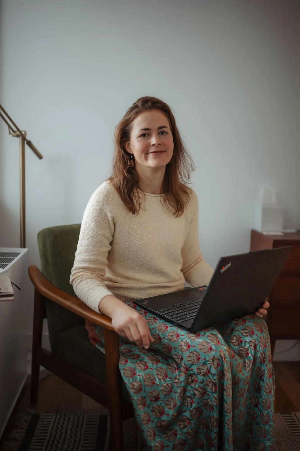 A woman with brown hair, wearing a cream sweater and colorful patterned skirt, sitting on a wooden chair with a laptop on her lap in a home office.