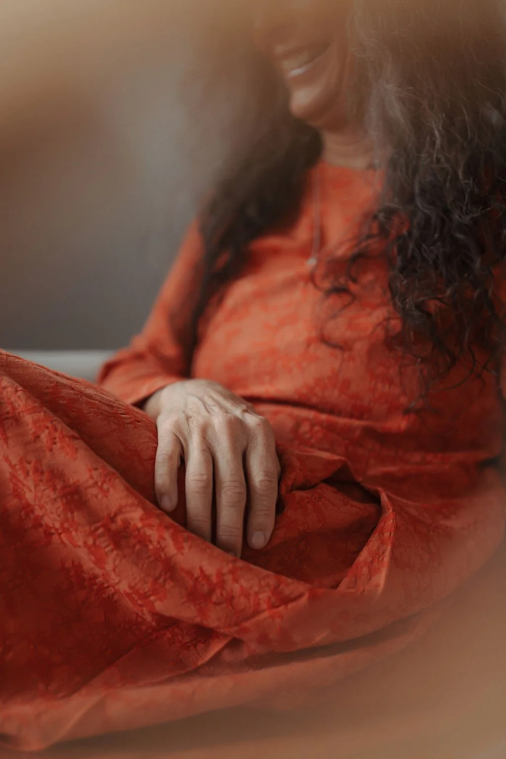 A woman dressed in a red/orange traditional outfit sitting with her hand resting on her lap, smiling and looking down.