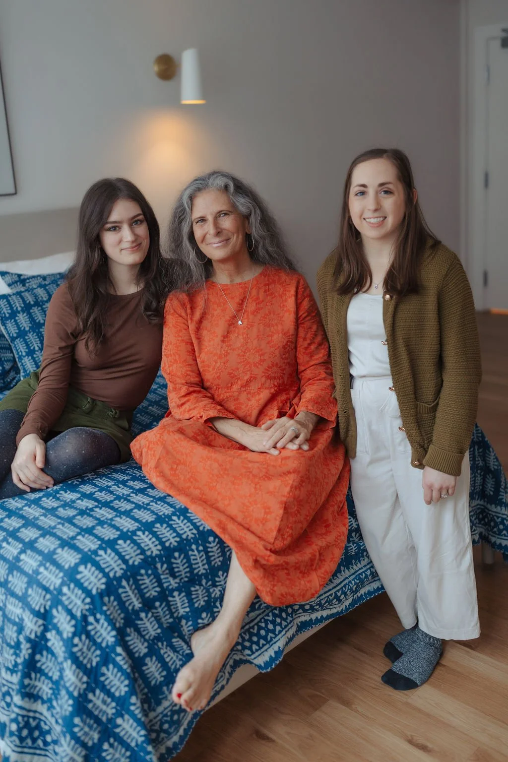 Three women sitting and standing on a bed in a bedroom, smiling at the camera. The woman in the middle has gray hair and is wearing an orange dress, while the other two women have brown hair and are wearing casual clothes.