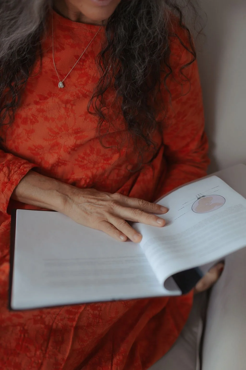 A woman with long, curly gray hair wearing a red-orange shirt with a floral pattern, holding an open book with a diagram of a human uterus on the page.