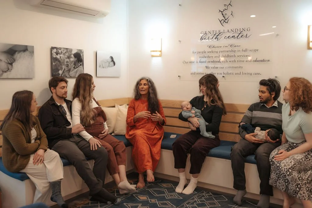 A diverse group of people sitting on a bench inside a birthing center, engaging in a discussion, with some holding infants, and framed photographs on the wall behind them.