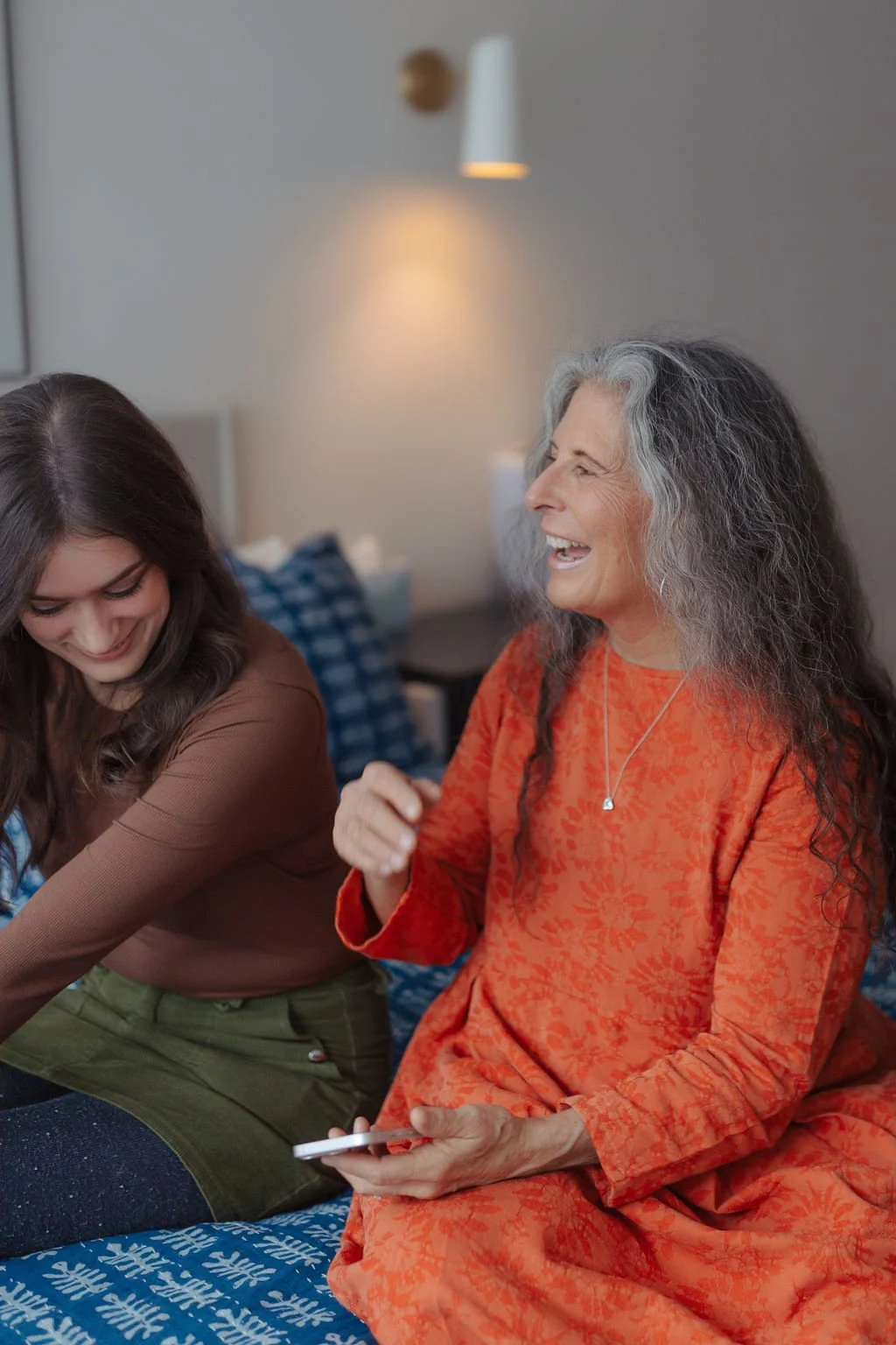 An elderly woman and a young woman are sitting on a bed, laughing and looking down at a smartphone.