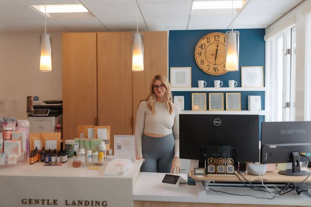 A woman standing behind a reception desk at a wellness or health clinic, with herbal remedies, essential oils, and supplements on display, near a blue wall with framed certificates, cups, and a large wall clock.