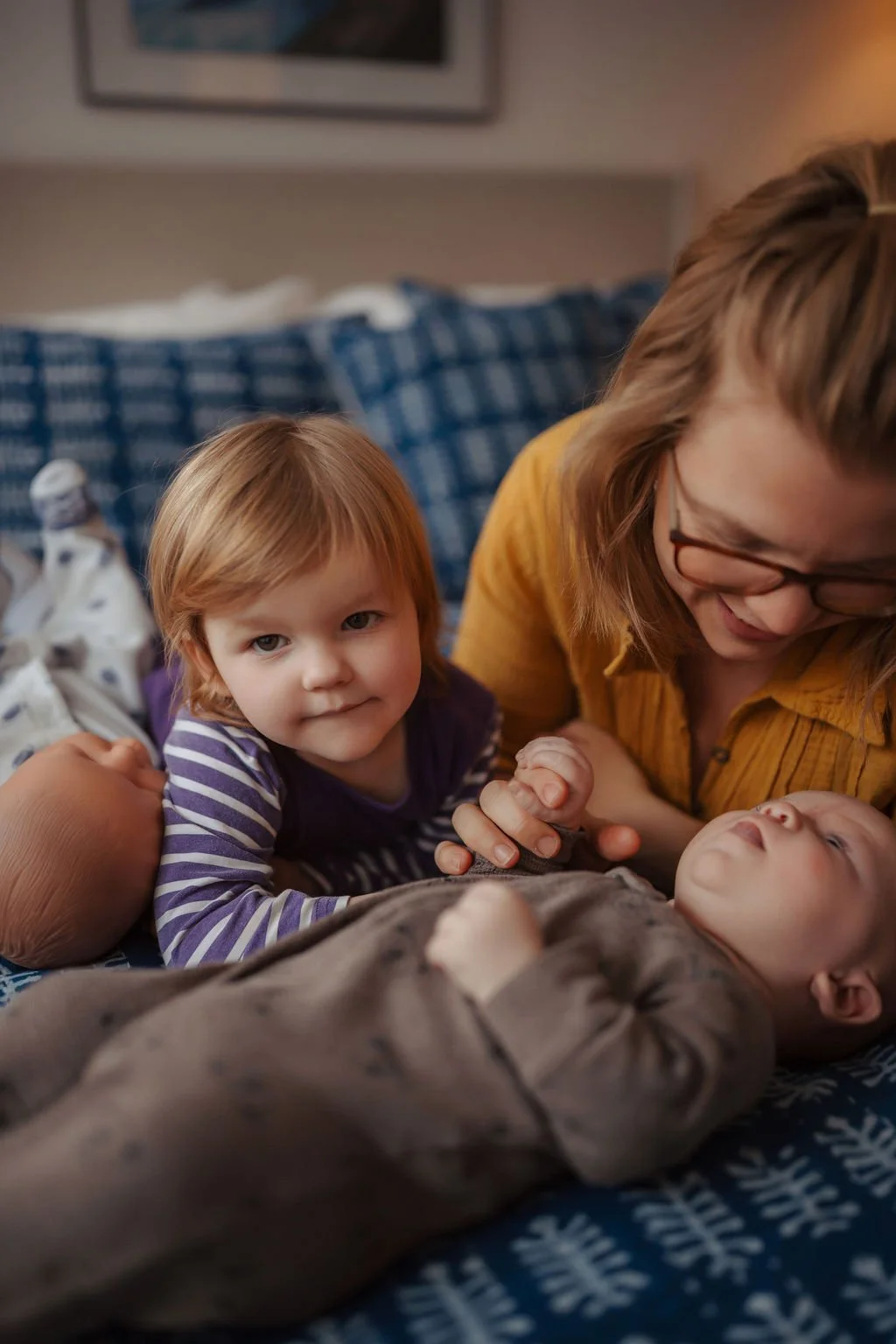 A woman with glasses and a young girl with red hair lie on a bed, smiling and holding hands with a baby laying on the bed.
