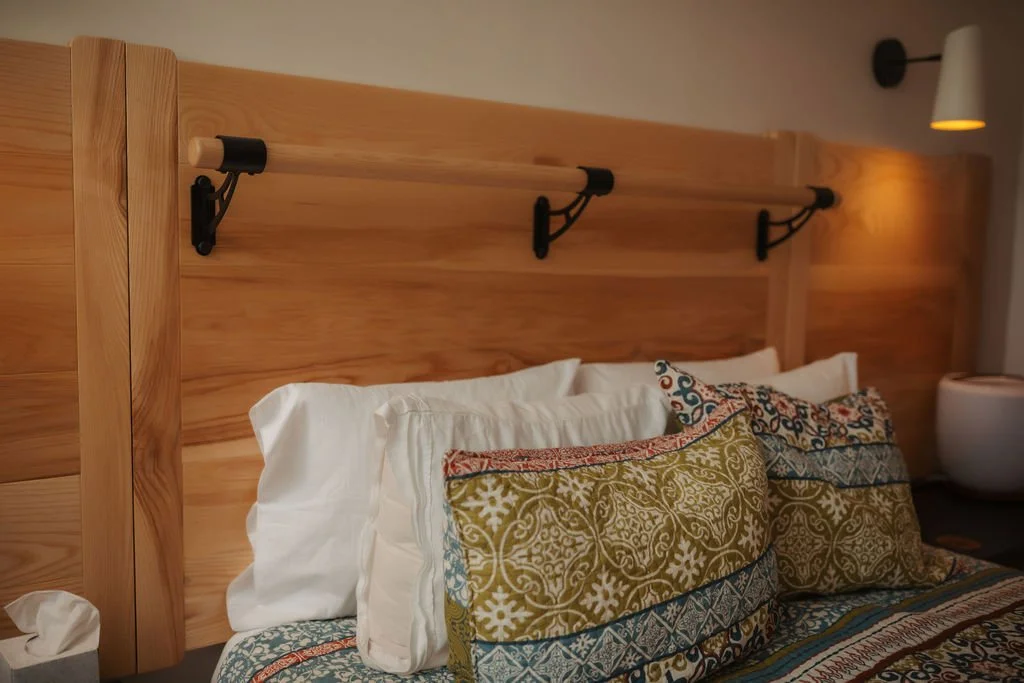 Close-up of a bed with colorful patterned pillows and a wooden headboard in a hotel room, with mounted reading lights and a modern bedside lamp.
