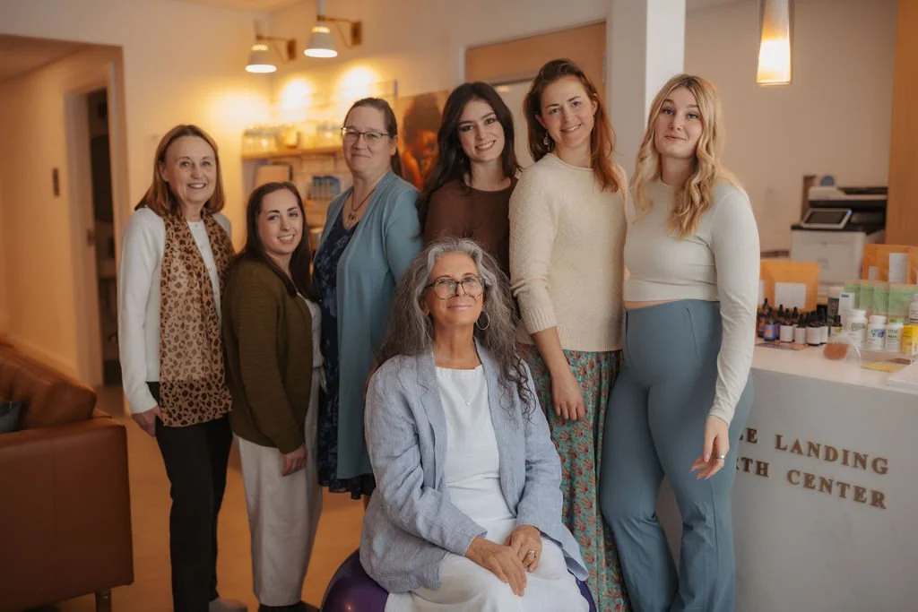 Seven women standing and sitting together in a warmly lit room, smiling, with a reception area and various bottles of medicine or skincare products on a counter in the background.