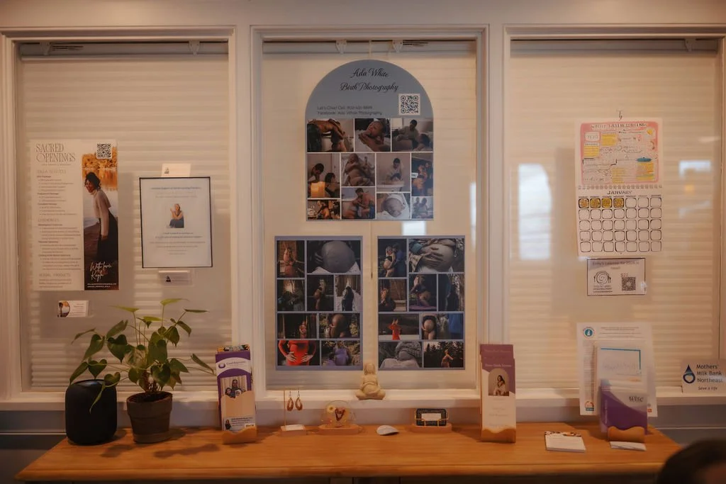 An information display board with photos and notices at a community center or office. Contains a large poster of a woman and the words "Sacred Openings," a smaller framed note, a calendar, brochures, and various small decorative items and a potted pl