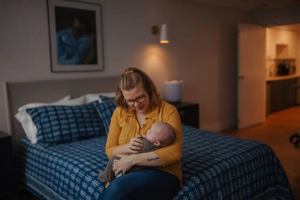 A woman with glasses and a yellow shirt holding a baby in a bedroom with a blue checkered bedspread, a picture on the wall, and warm lighting in the background.