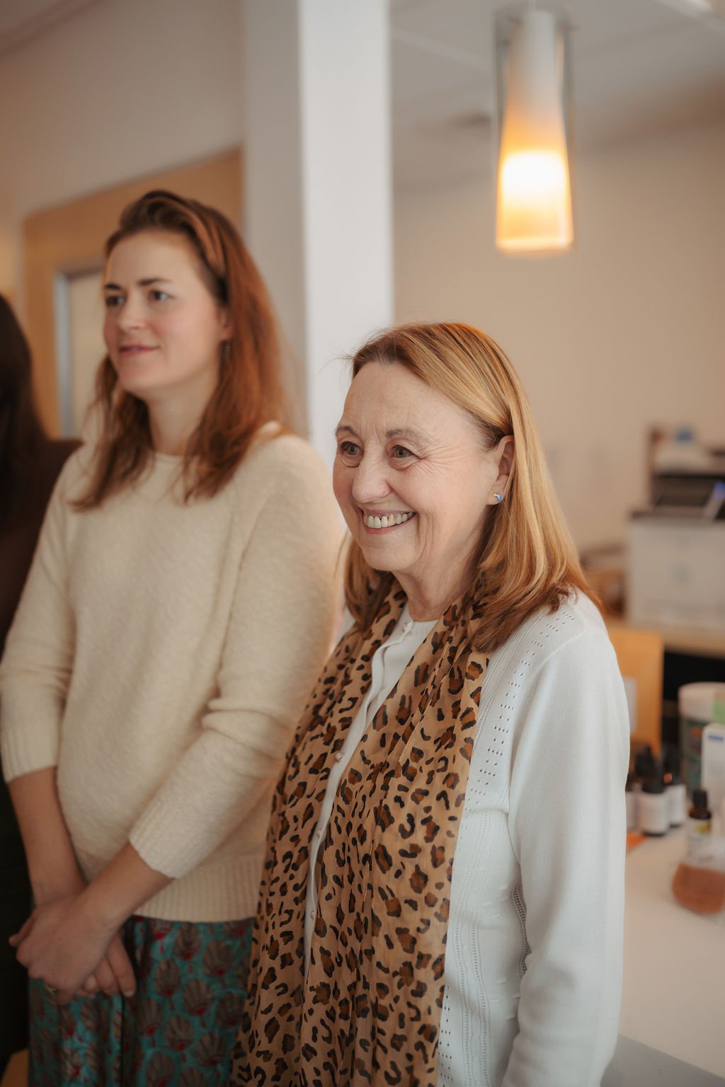 Two women standing indoors, smiling. The woman on the right has red hair, wearing a white sweater and a leopard print scarf. The woman on the left has brown hair, wearing a cream sweater.
