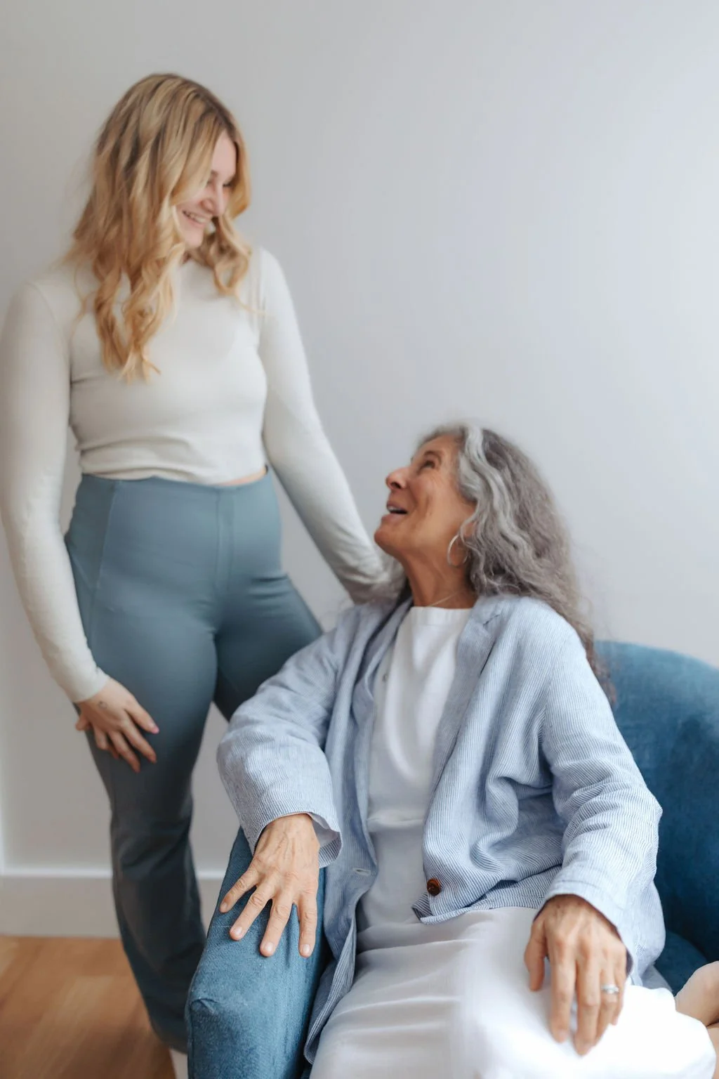 A young woman standing next to an elderly woman sitting in a chair, smiling and looking at each other, in a bright room with a plain background.