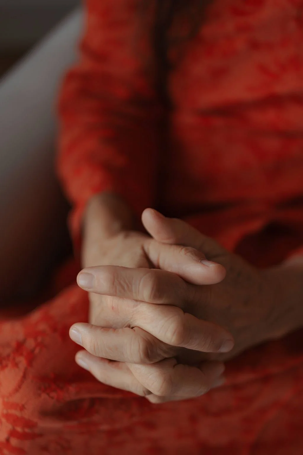 Close-up of an elderly person's hands clasped together, resting on a red patterned fabric.
