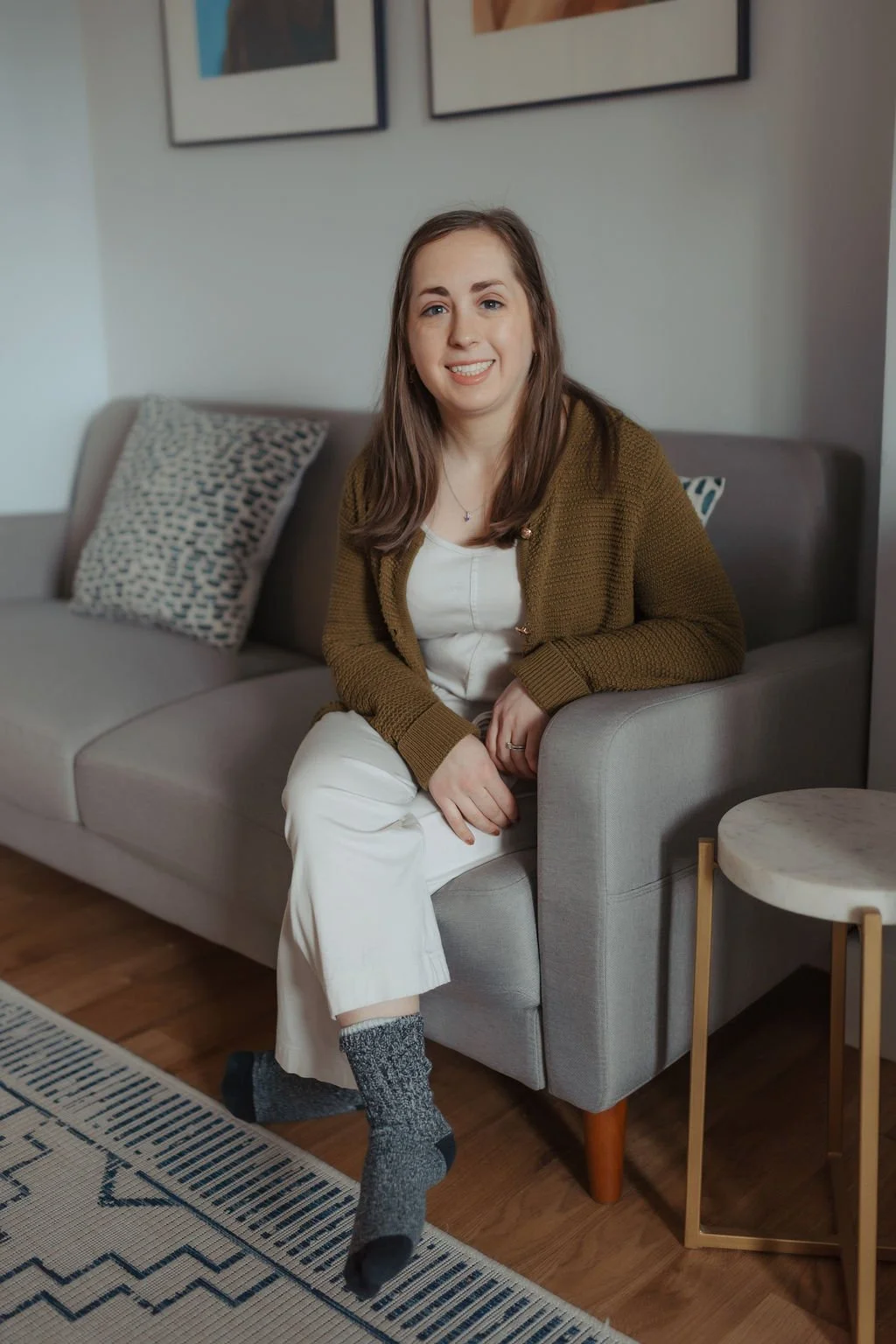 A woman sitting on a gray sofa in a living room, smiling at the camera, wearing a brown cardigan, white top and pants, with gray socks. A patterned pillow is on the sofa, with framed pictures on the wall above.