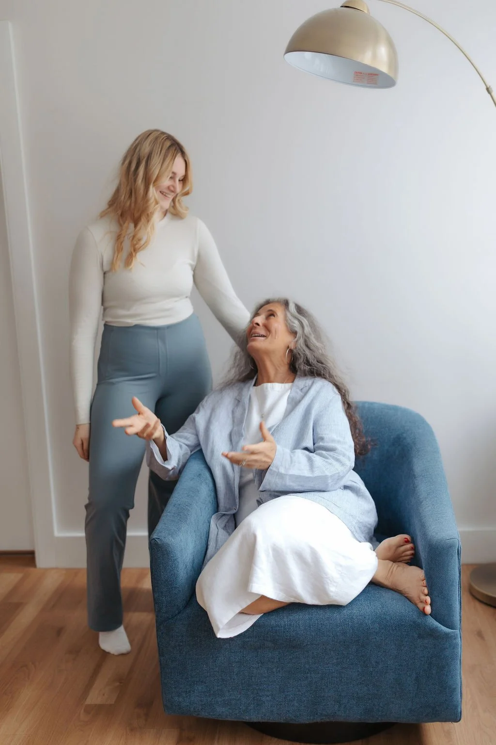 An elderly woman with gray hair sitting in a blue armchair, smiling and talking, while a younger woman with long blonde hair and light-colored clothing stands beside her, smiling and touching her head, in a cozy room with a modern floor lamp and wood