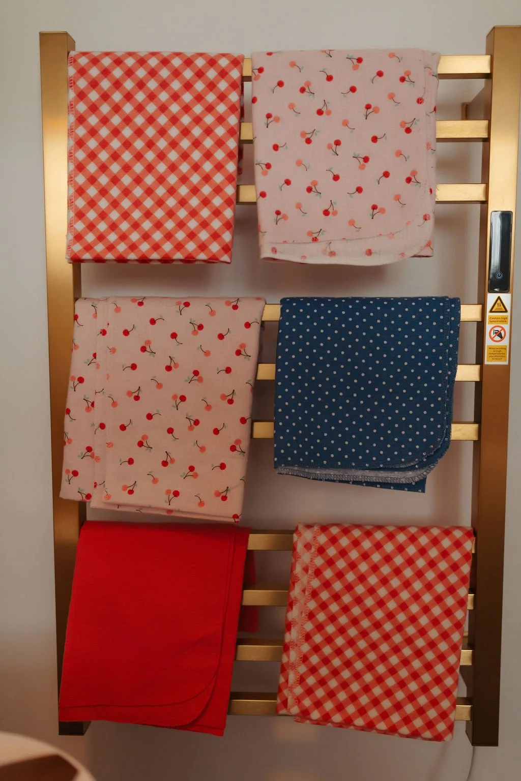 Six folded cloth napkins with various patterns, displayed on a metallic rack. The patterns include red and white checkered, cherry print on light pink, navy blue with white polka dots, and solid red.