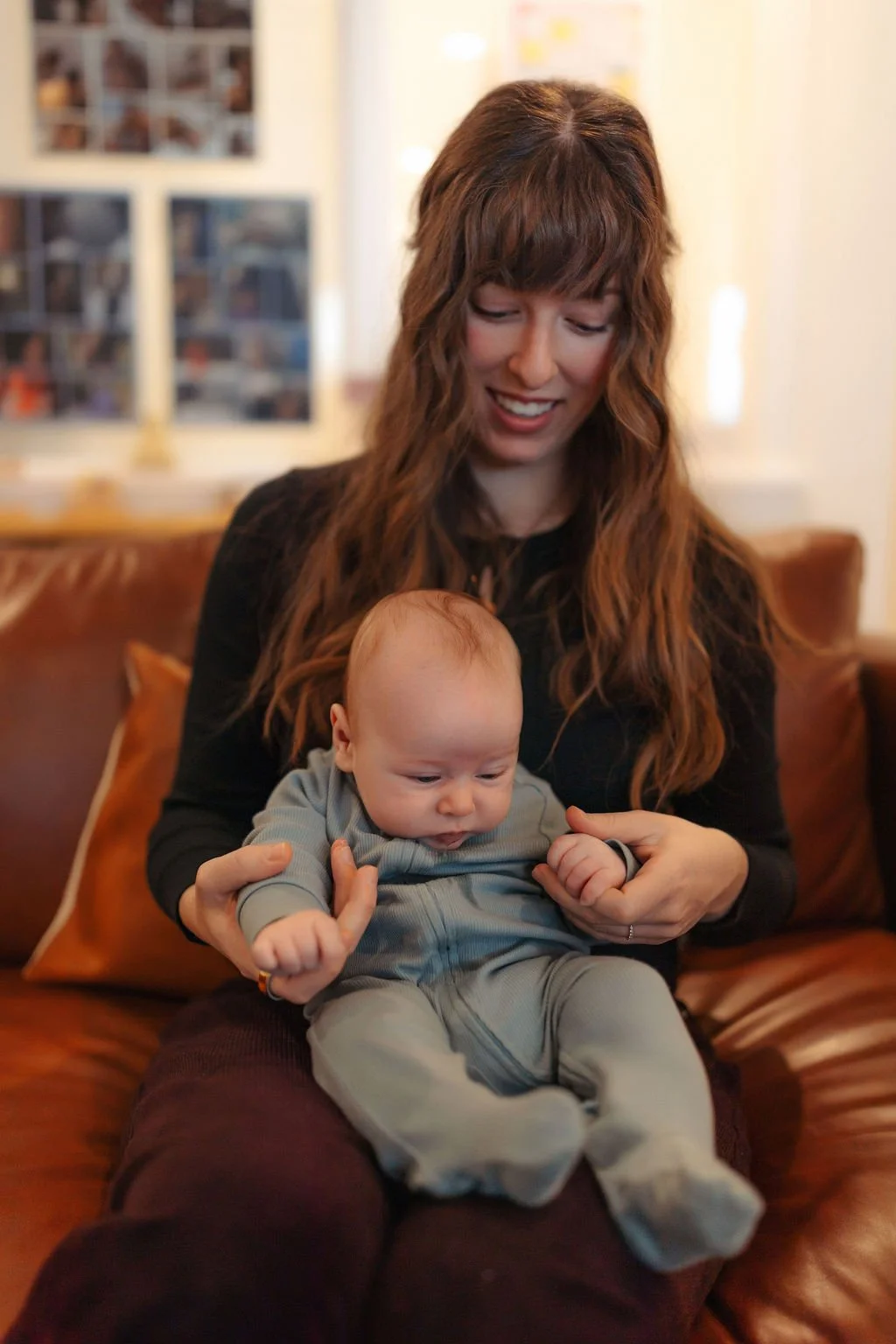A woman with long wavy brown hair smiling while holding a baby dressed in gray, sitting on a leather couch in a cozy room with photos on the wall.