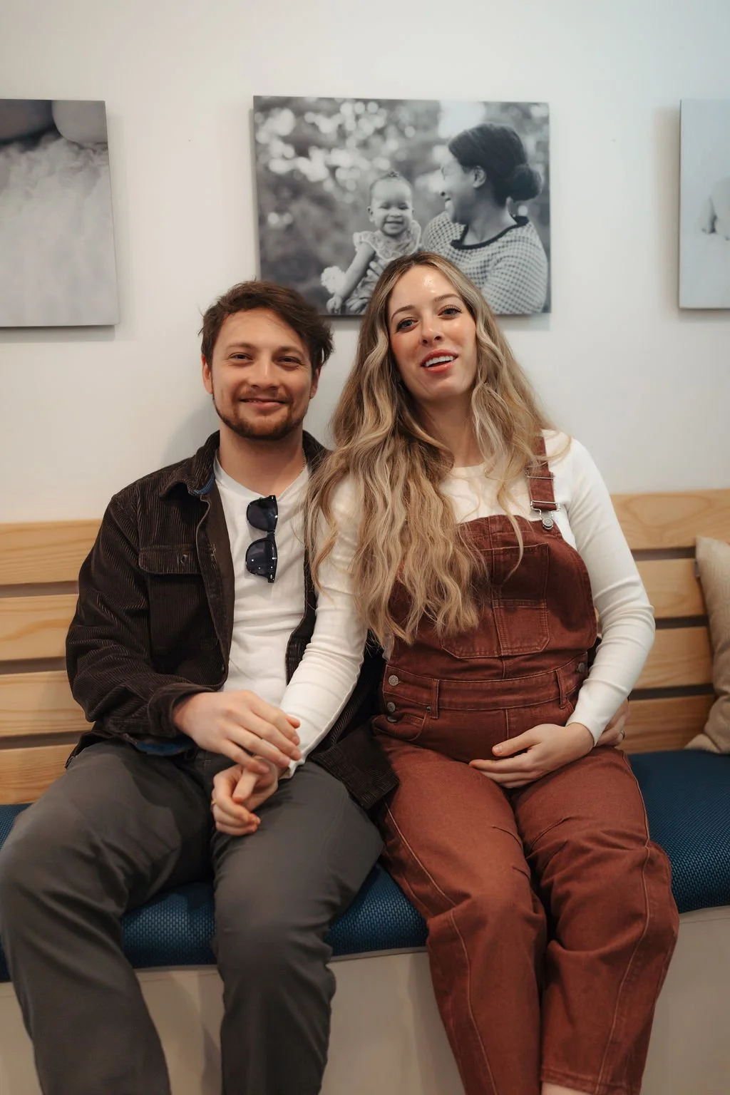 A smiling man and pregnant woman sitting on a blue cushioned bench, holding hands, with black and white family photos hanging on the white wall behind them.