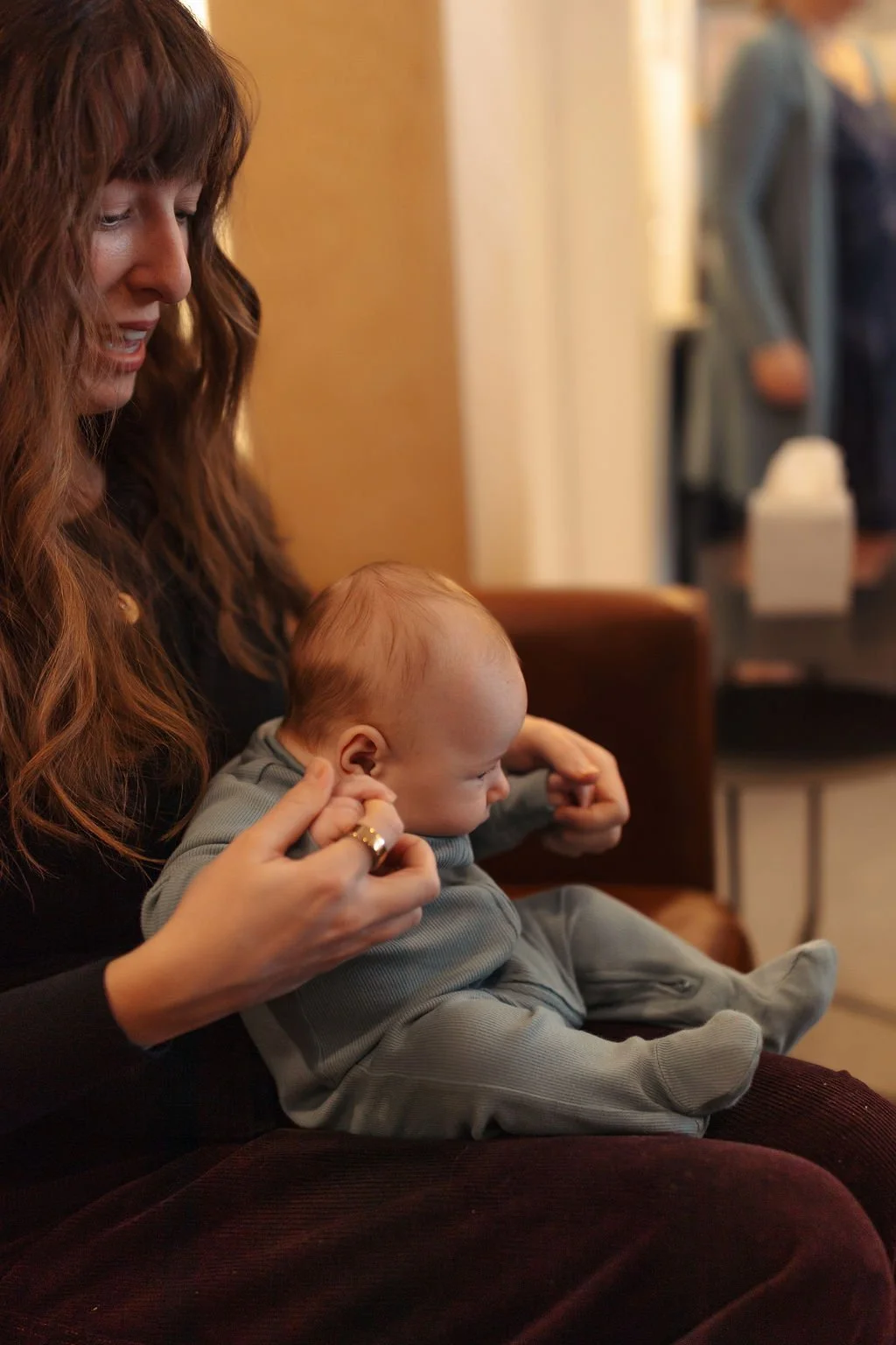 A woman with long wavy red hair is sitting on a brown chair holding a baby with light hair and fair skin on her lap. The woman is smiling slightly while the baby appears to be looking down at something. The background is an indoor setting with blurre