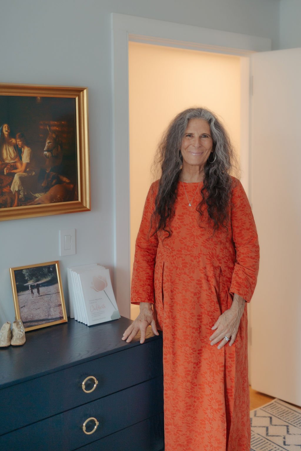 A woman with long, curly gray hair, wearing an orange patterned dress, stands beside a dark dresser with decorative rings on the drawers. There are picture frames, pamphlets, and a pair of shoes on the dresser. Behind her is a white door and a framed