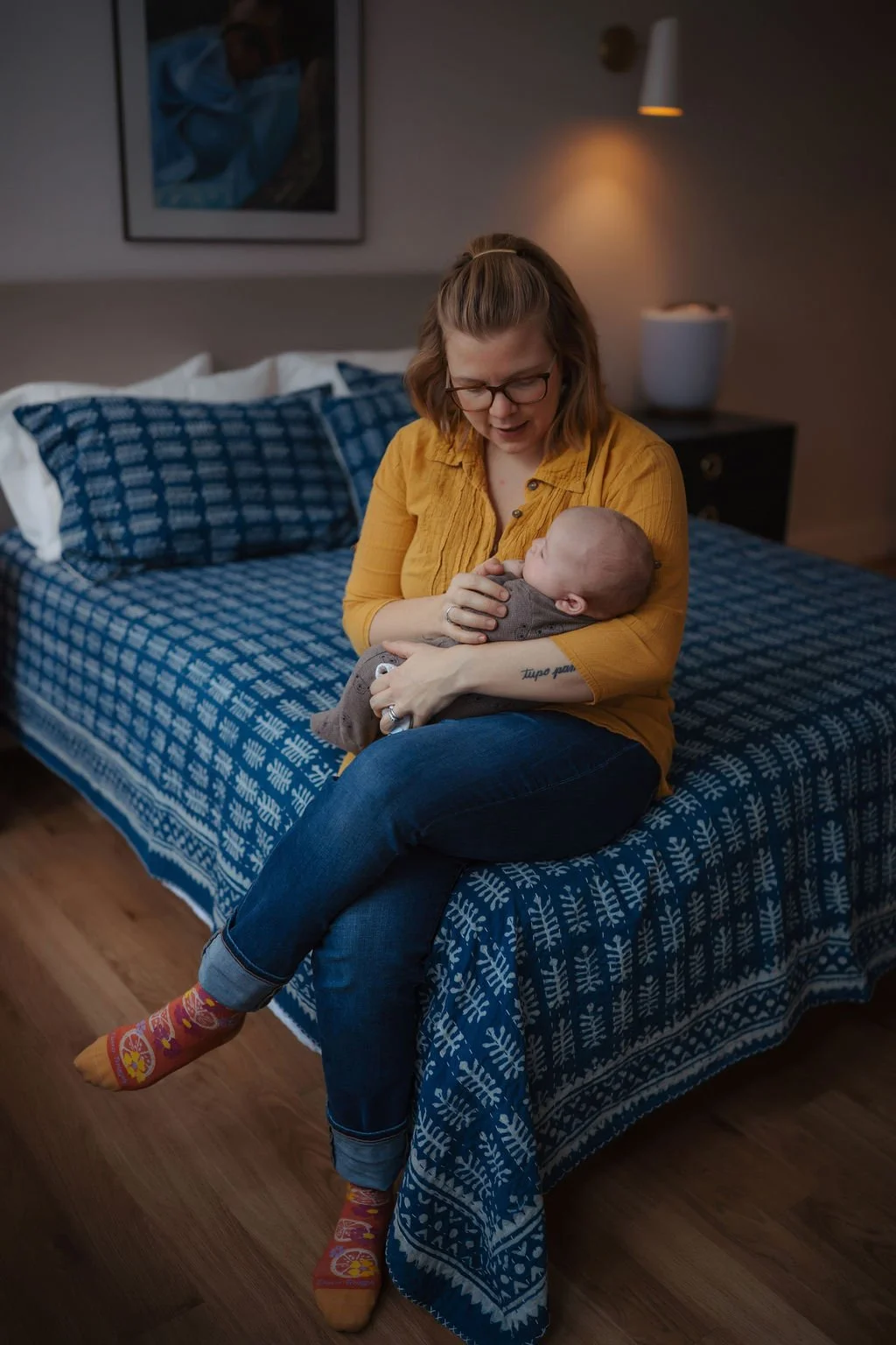 A woman with glasses and a yellow shirt sits on a bed holding a baby wrapped in a brown blanket, looking at the baby with a smile in a dimly lit bedroom with a blue patterned bedspread.