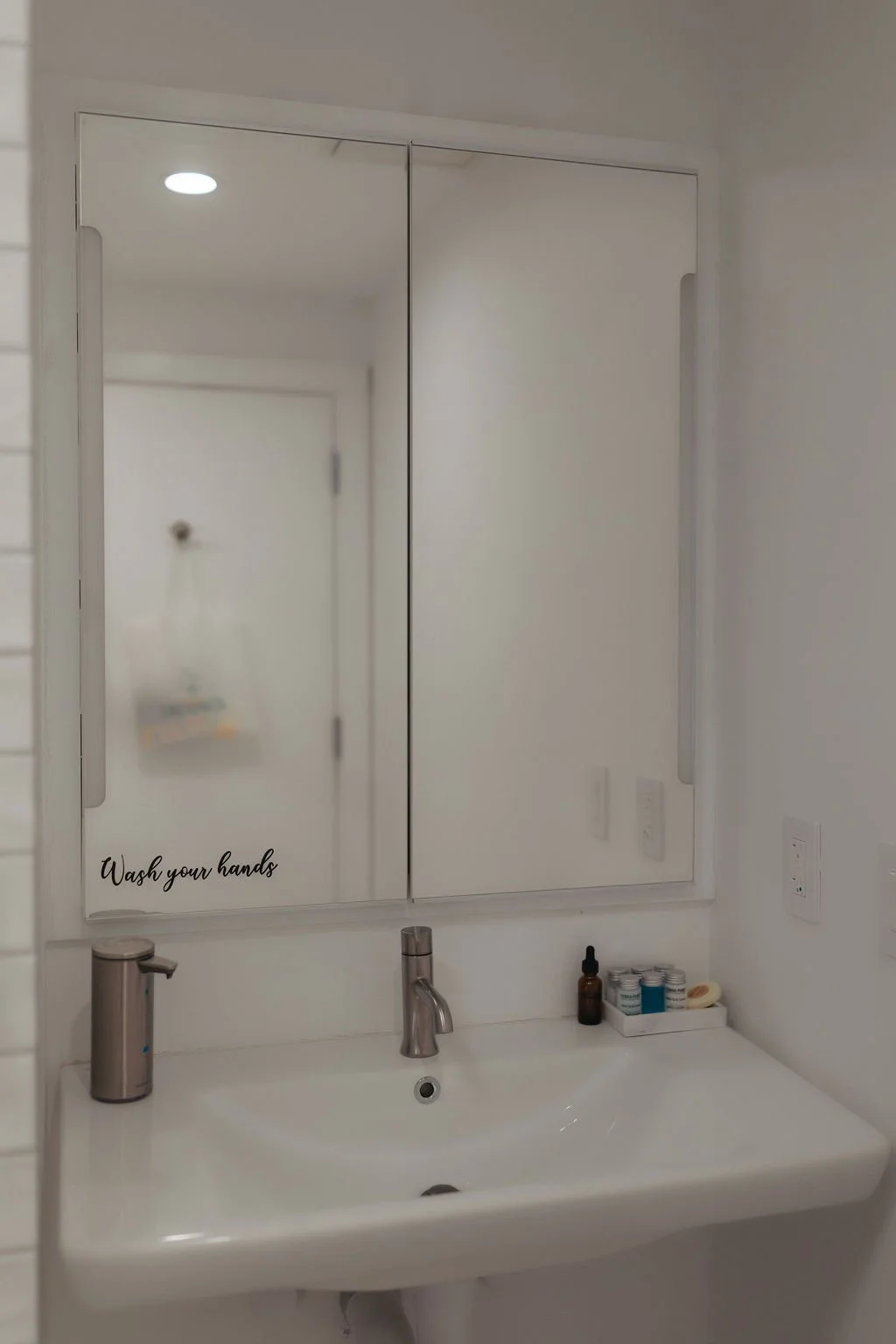 Bathroom mirror with the words "Wash your hands" written on the lower left corner, sink, faucet, hand soap dispenser, brown dropper bottle, small containers, and a small tray with skincare products.