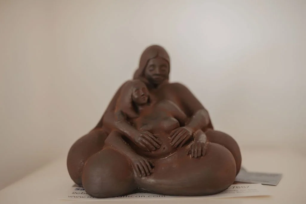 A sculpture of a woman with two children on her lap, with detailed facial expressions and body features, made of dark material, displayed on a surface against a plain wall.