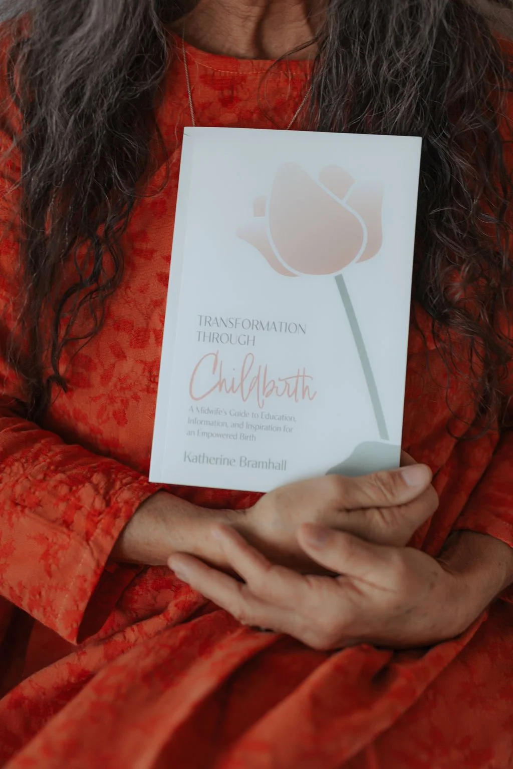 A person wearing a red textured dress holding a book titled 'Transformation Through Childbirth' by Katherine Bramhall, with long dark curly hair.
