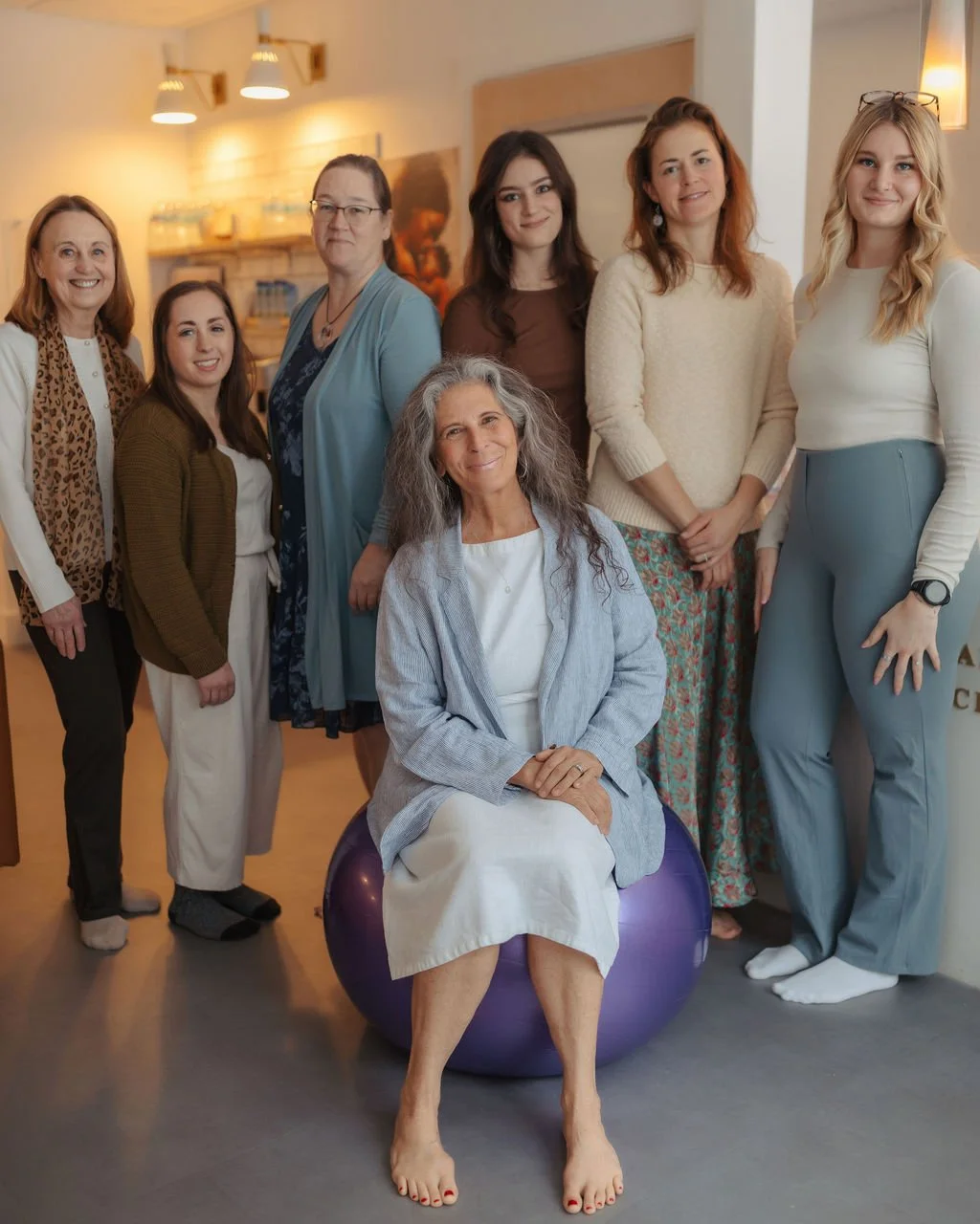 Group of women of various ages standing in a cozy room, with a woman sitting on a purple exercise ball in front, all smiling at the camera.