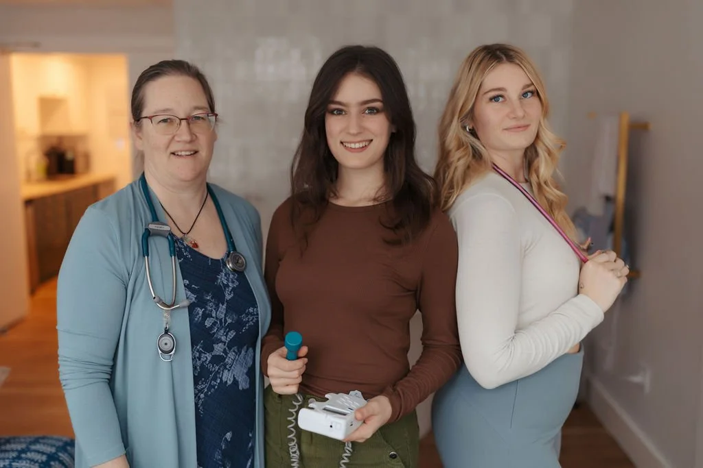 Three women standing together indoors, smiling at the camera. One is a healthcare professional wearing a blue coat with a stethoscope around her neck, and the other two women are holding what appear to be medical or healthcare tools.