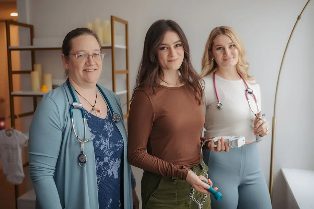 Three women standing together inside a healthcare setting. The woman on the left is a healthcare professional wearing a light blue cardigan and stethoscope. The woman in the middle is a young patient holding a blood pressure cuff and a small medical 