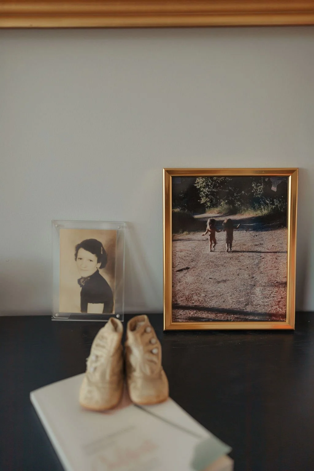 A desk with a pair of beige sneakers and an open book in the foreground. On the wall behind, a vintage black and white portrait of a woman in a dark dress on the left and a framed photograph of two children walking on a dirt path surrounded by trees 
