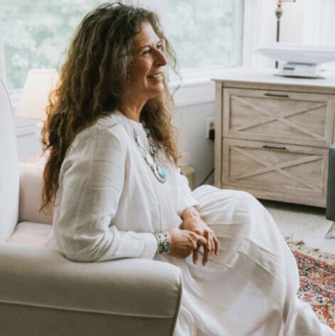 Smiling woman with long, wavy hair sitting on a white couch in a bright room, with a wooden dresser and a lamp in the background.
