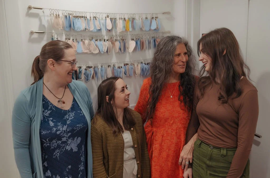 Four women standing together indoors, engaged in conversation and smiling. Behind them, a wall decorated with multiple rows of clothespins holding small sock-like objects in various pastel colors.