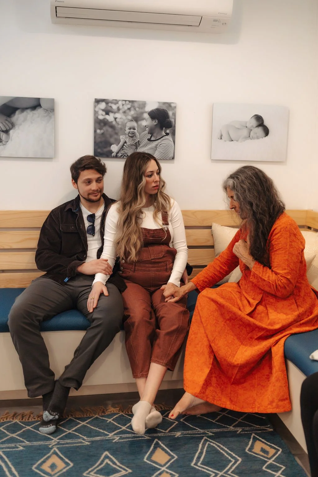 Three people sitting on a sofa, with a young man on the left, a young woman in the middle, and an older woman on the right dressed in orange. The older woman is holding the young woman's hand, and they are sitting in a cozy room with framed black-and