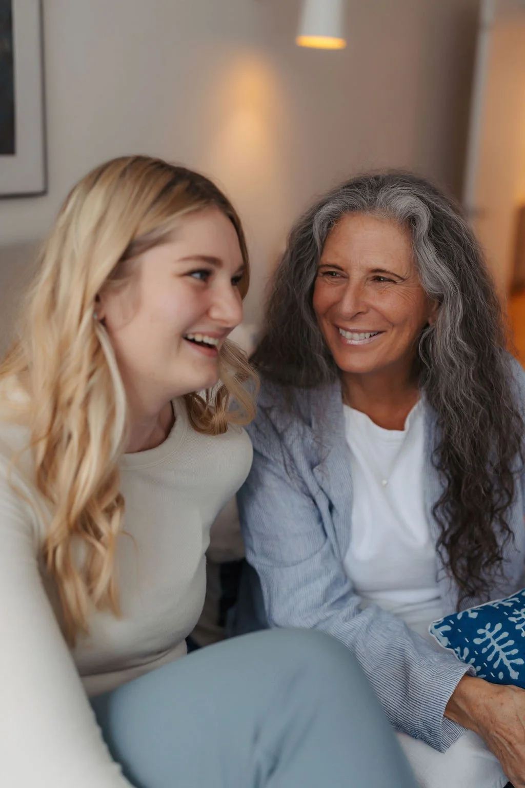 A young woman and an older woman with gray hair smiling and looking at each other, sitting in a cozy indoor setting.