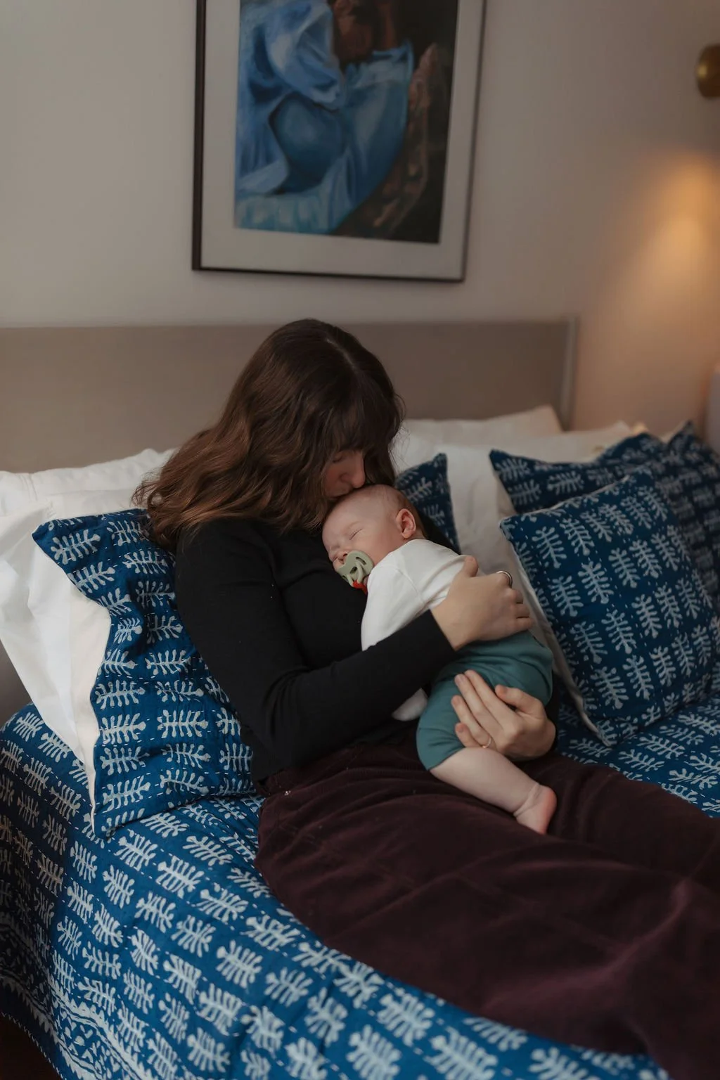 A woman with brown hair, wearing a black long-sleeve shirt, sits on a bed holding a sleeping baby with a pacifier in a white shirt and teal pants. The bed has blue patterned pillows and a duvet with similar pattern. There is a framed abstract painting hanging on the wall in the background that depicts a person in blue.