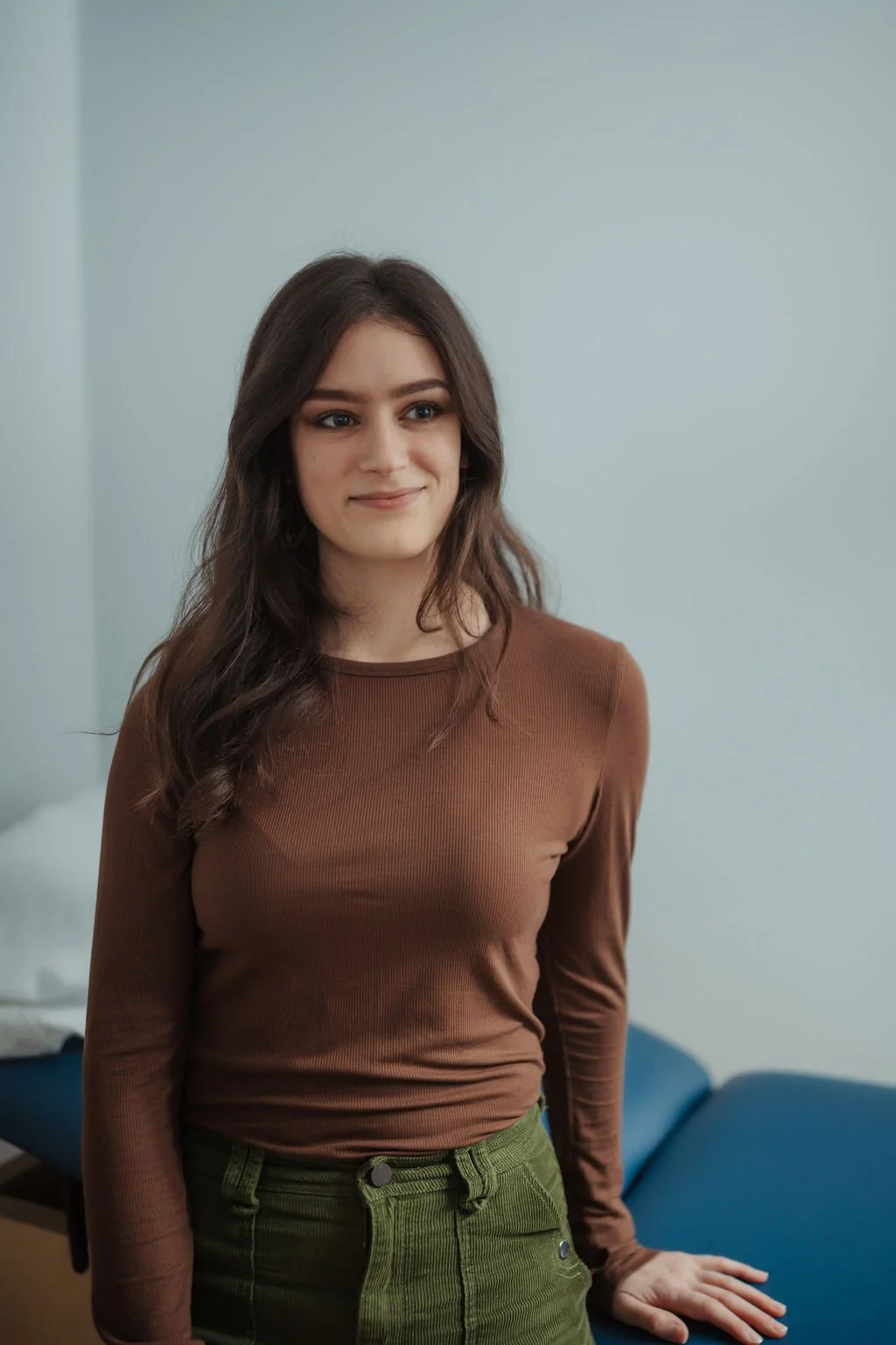 A young woman with long brown hair standing in a room with a light blue wall, wearing a brown long-sleeve top and green pants.