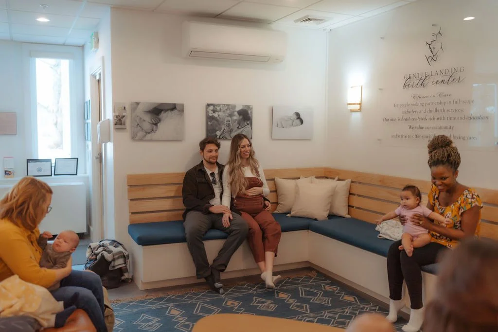 Waiting room at a birth center with people and babies, including a woman holding a baby and a woman with a baby on her lap, on a blue and beige patterned rug.