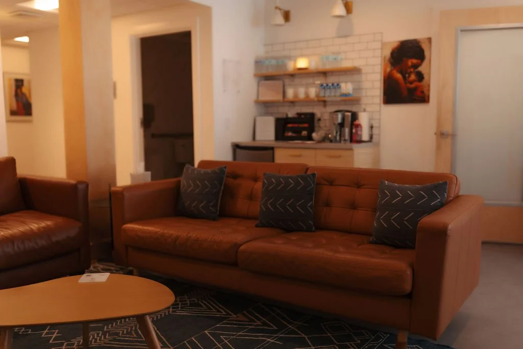 Living room with brown leather sofa and armchair, black pillows with white arrow patterns, wooden coffee table, and a kitchenette with shelves, coffee maker, and artwork on the wall.