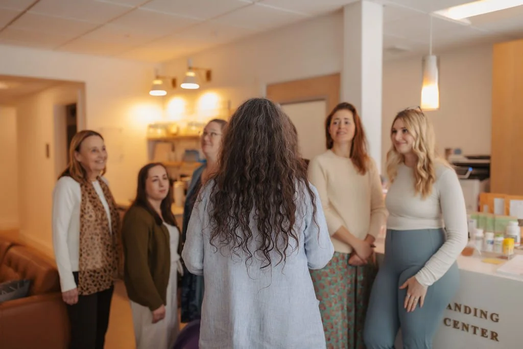 Group of five women engaging in conversation in a cozy indoor setting, with a countertop displaying various bottles and a sign that reads 'ANDING CENTER'.