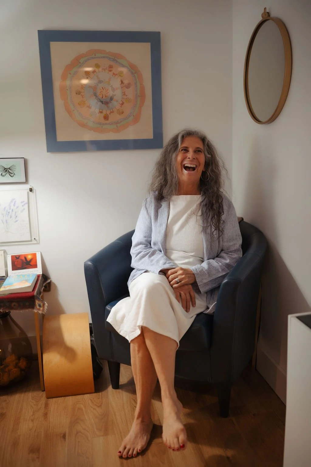A woman with long gray hair sitting on a black armchair, laughing joyfully, in a room with framed artwork on the wall, a mirror, and a side table with books, a photo, and decorative items.