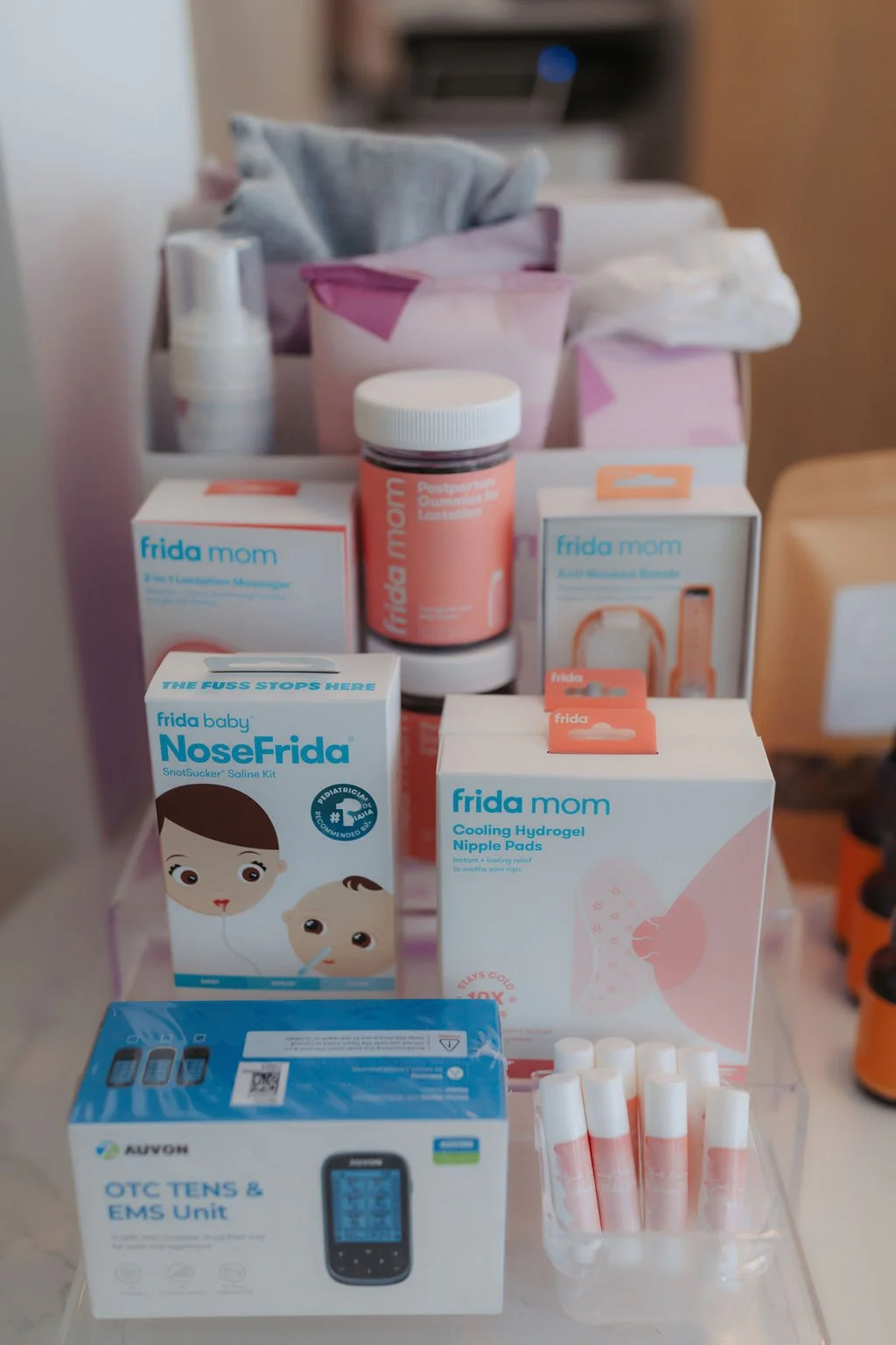 A collection of maternity and baby care products including nasal snot sucker, cooling hydrogel nipple pads, postpartum gummies, anti-nausea bands, OTC TENS and EMS unit, lip balms, and tissue packs on a table.