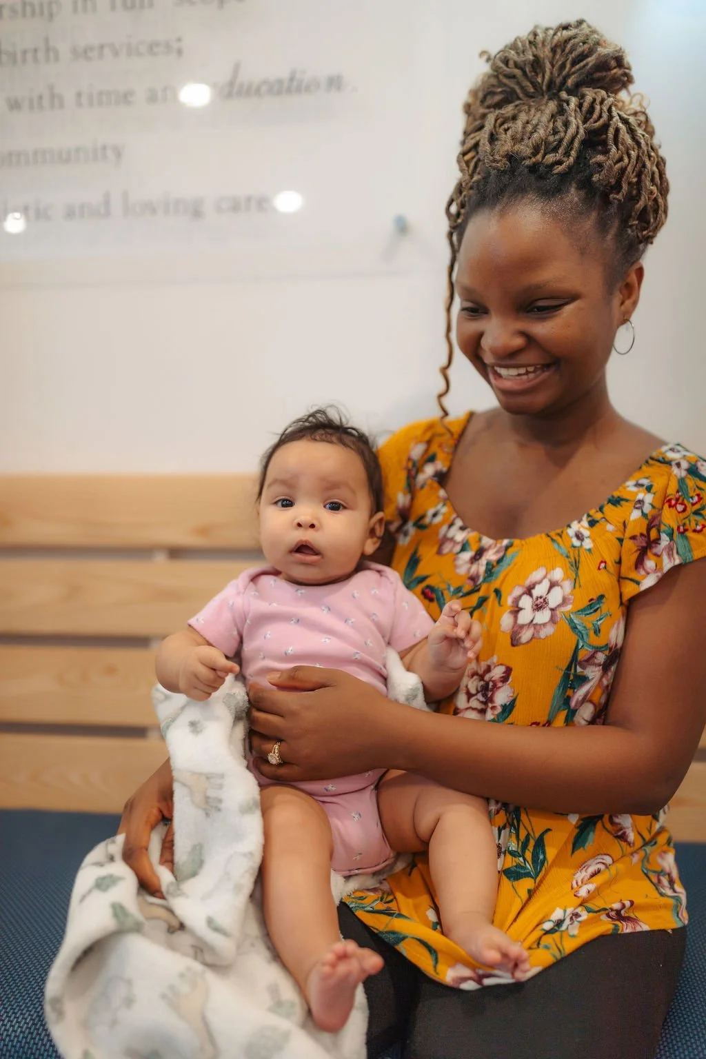 A woman with braided hairstyle and hoop earrings holding a young baby girl in her lap. The woman is smiling, and the baby is looking at the camera. They are seated on a bench against a wall with a sign partially visible in the background.
