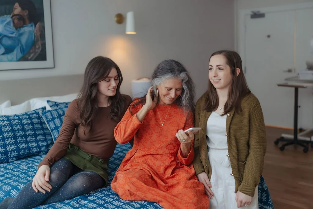 Three women sitting on a bed, smiling and looking at a smartphone.