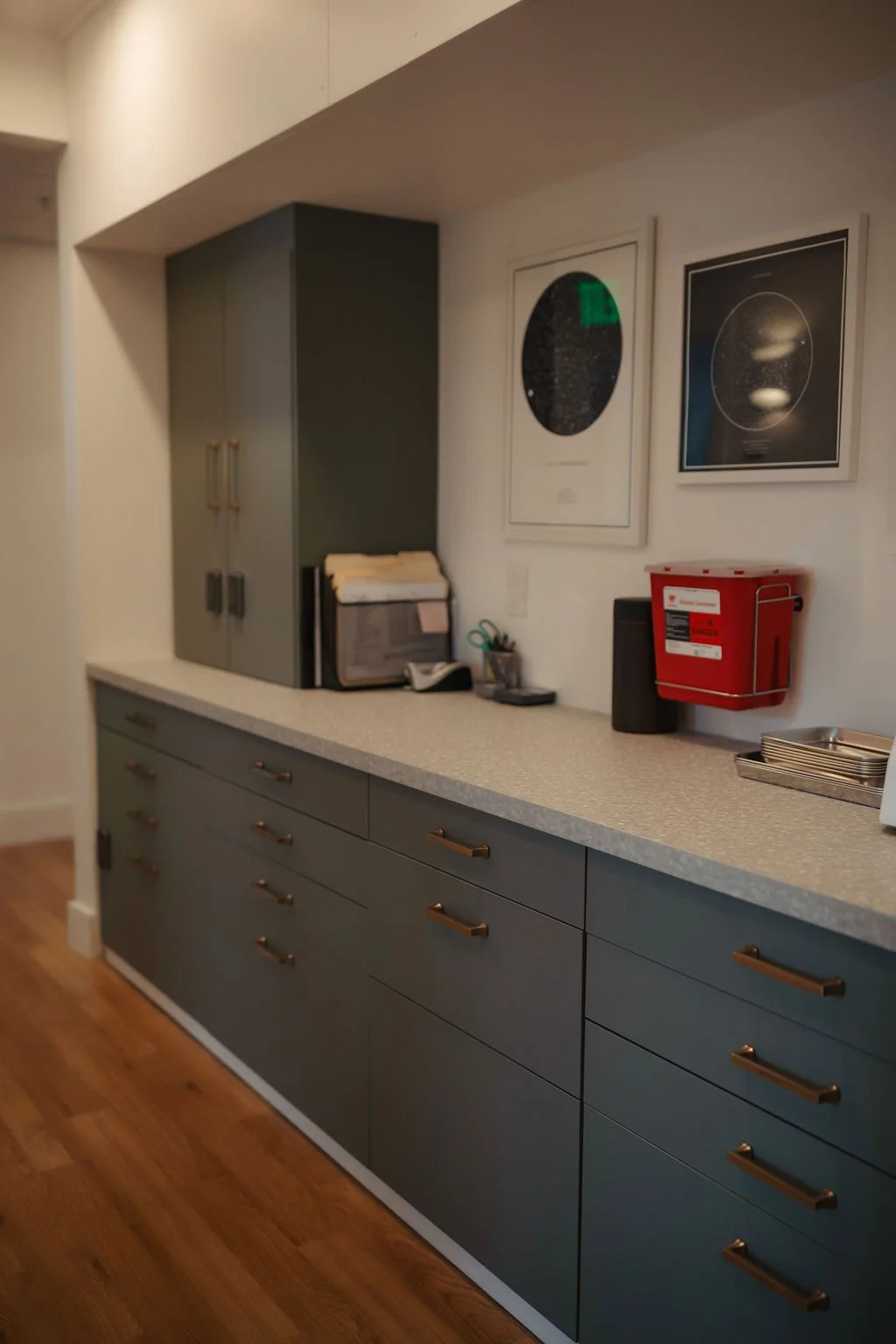 A modern kitchen countertop with gray cabinets and gold handles, a framed space-themed artwork on the wall, a red fire safety box, and various office supplies.